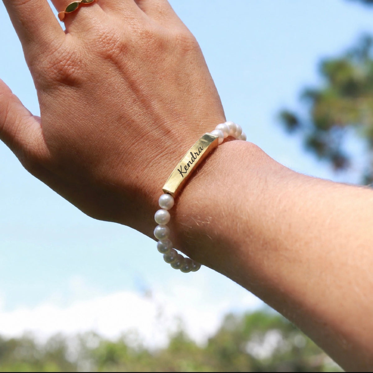 A gold bar bracelet engraved with the name 'Kendra' worn on a person's wrist, paired with a white pearl beaded bracelet, shown outdoors against a blurred blue sky and green trees.