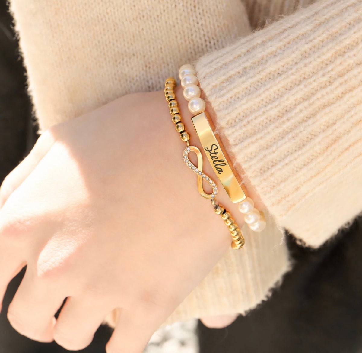 A close-up of a person's wrist wearing three bracelets: a gold bar engraved with 'Stella', a gold infinity symbol with small clear stones, and a pearl bead bracelet.