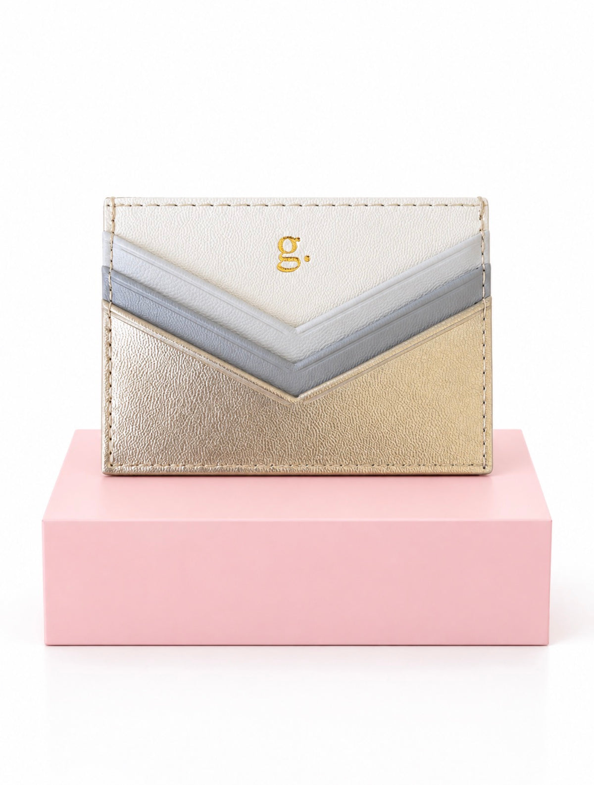 A chevron-patterned leather card holder with metallic gold, silver, and off-white panels, featuring a small gold 'g.' initial on the front, displayed on a pink block against a white background.