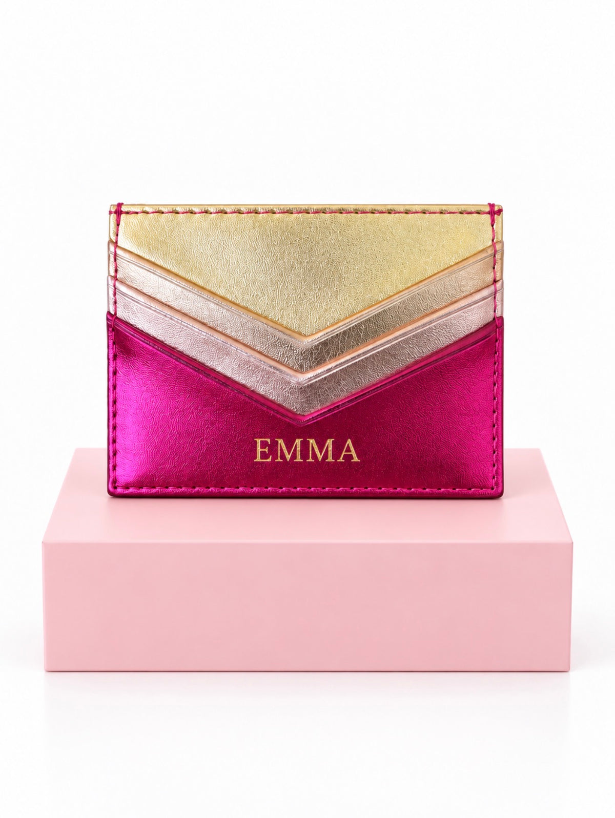 A metallic chevron card holder with a hot pink lower section, a rose gold middle section, and a gold top section, featuring the name 'EMMA' in gold lettering. It is displayed on a light pink block against a white background.