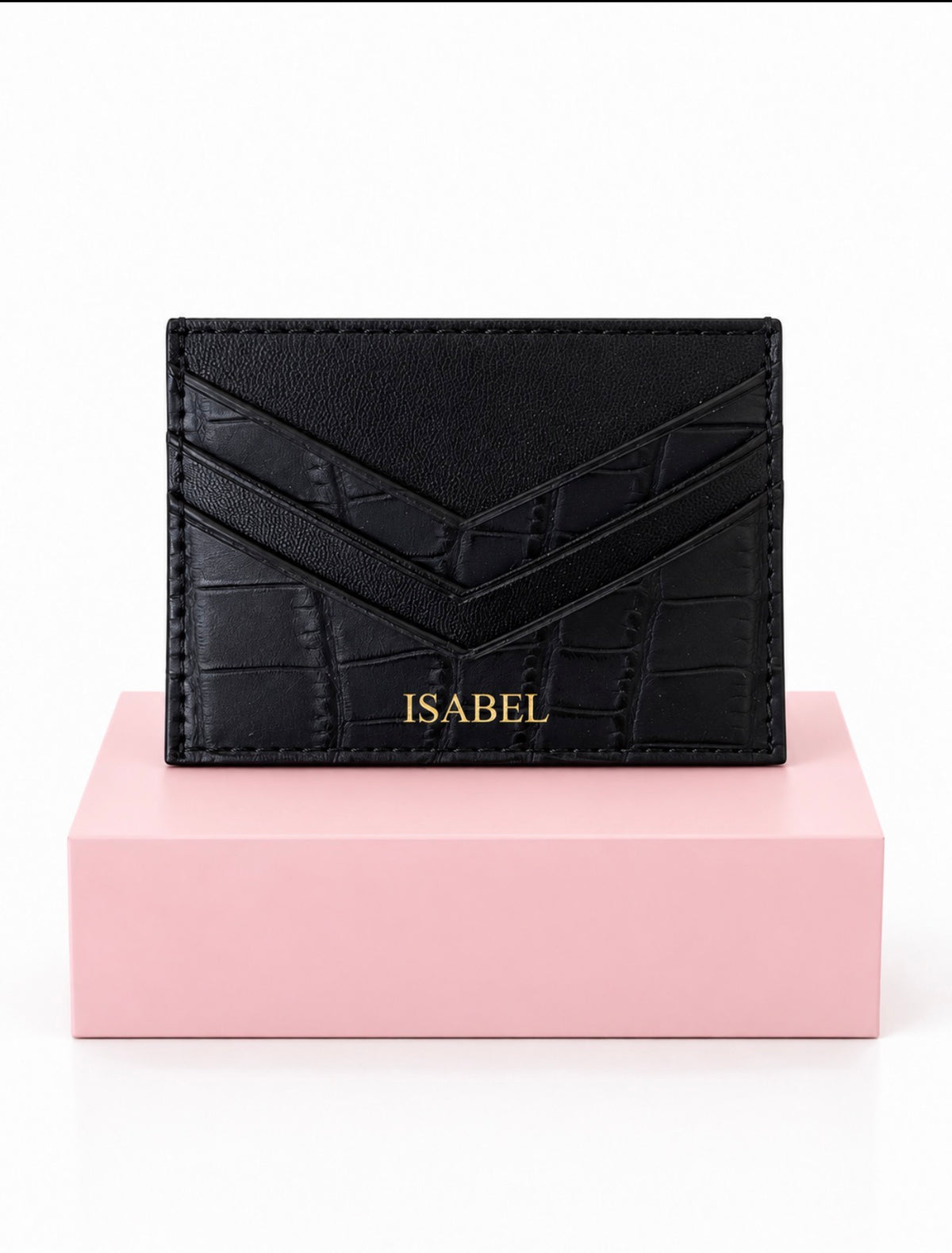A black faux-crocodile leather card holder with a gold 'ISABEL' name, displayed on a pink block against a white background.