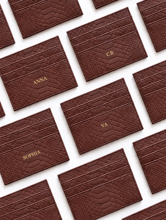 A close-up, top-down view of multiple dark brown textured leather cards arranged diagonally on a white background. Some cards are personalized with gold foil names including ANNA, CB, SOPHIA, and VA.