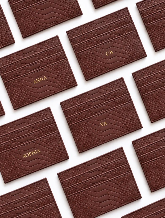 A close-up, top-down view of multiple dark brown textured leather cards arranged diagonally on a white background. Some cards are personalized with gold foil names including ANNA, CB, SOPHIA, and VA.