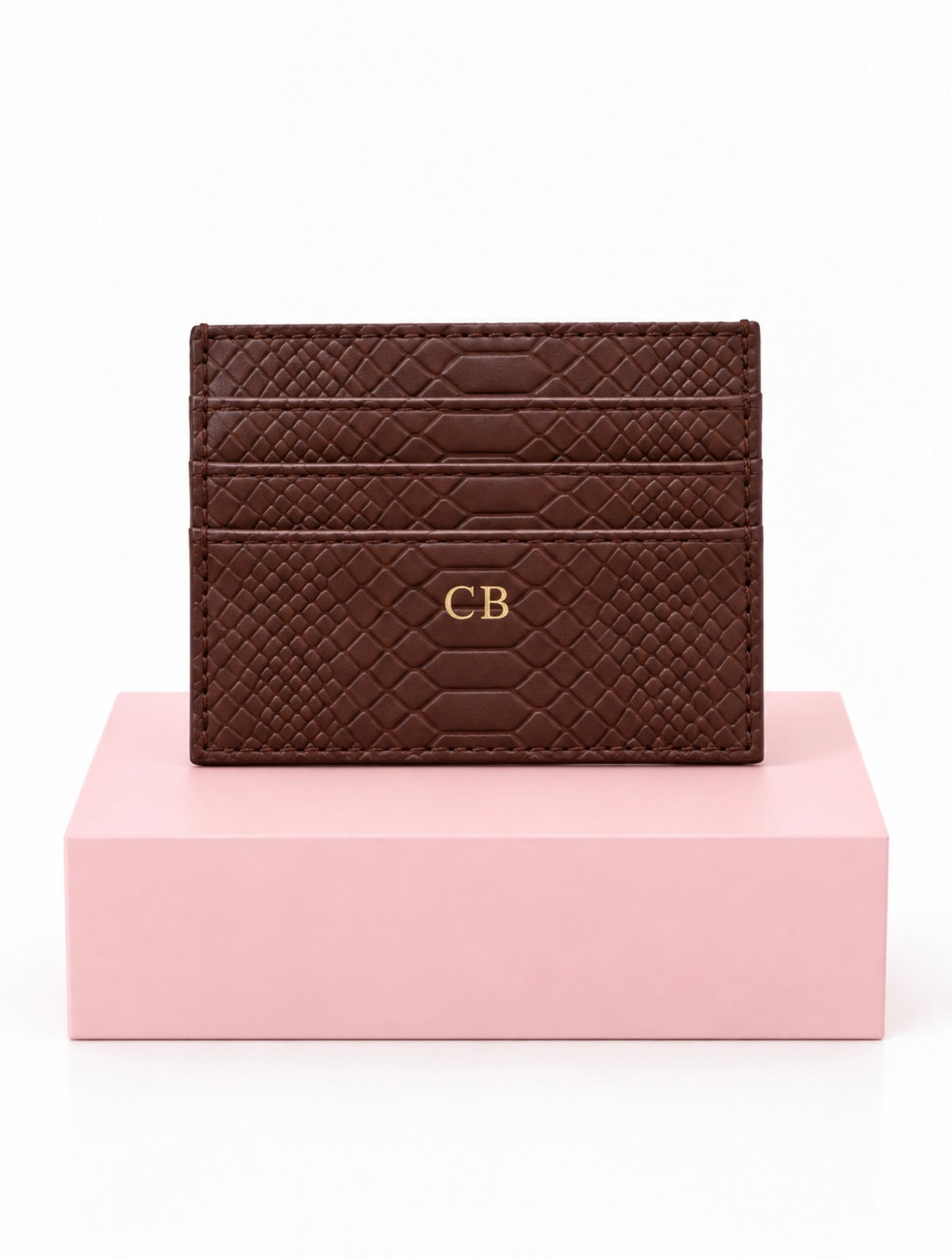 A dark brown textured card holder wallet with a gold 'CB' monogram, displayed on a light pink rectangular box against a white background.