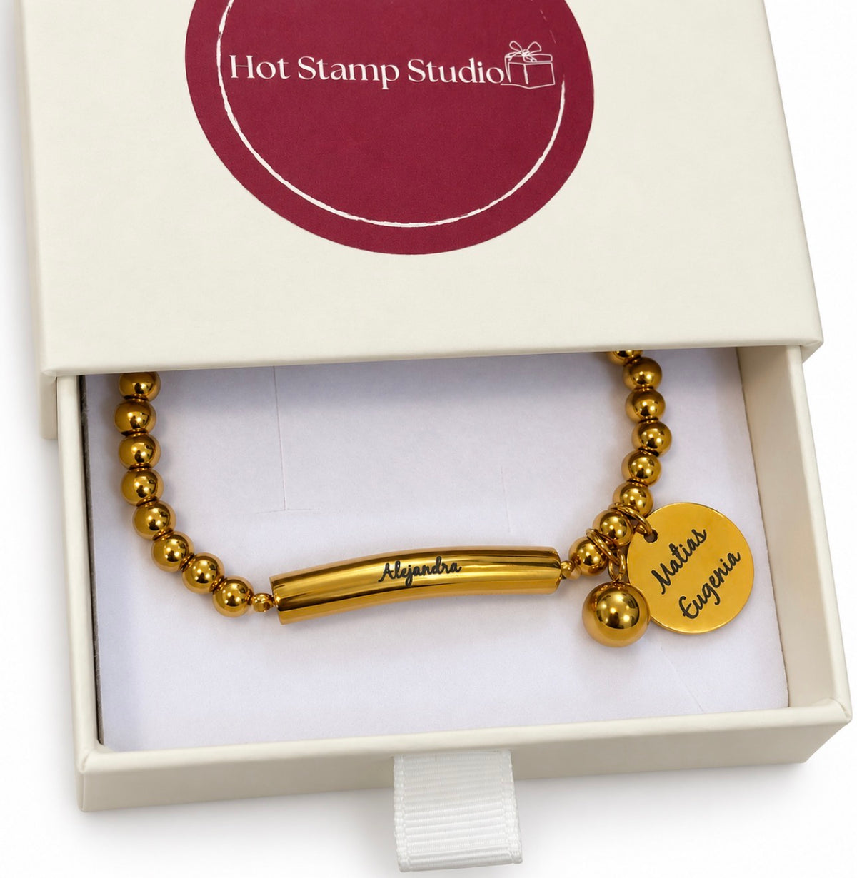A gold bracelet with the name Alejandra written across a bar. It includes a circular charm with the names Matias and Eugenia in cursive. The bracelet is sitting inside a gift box 